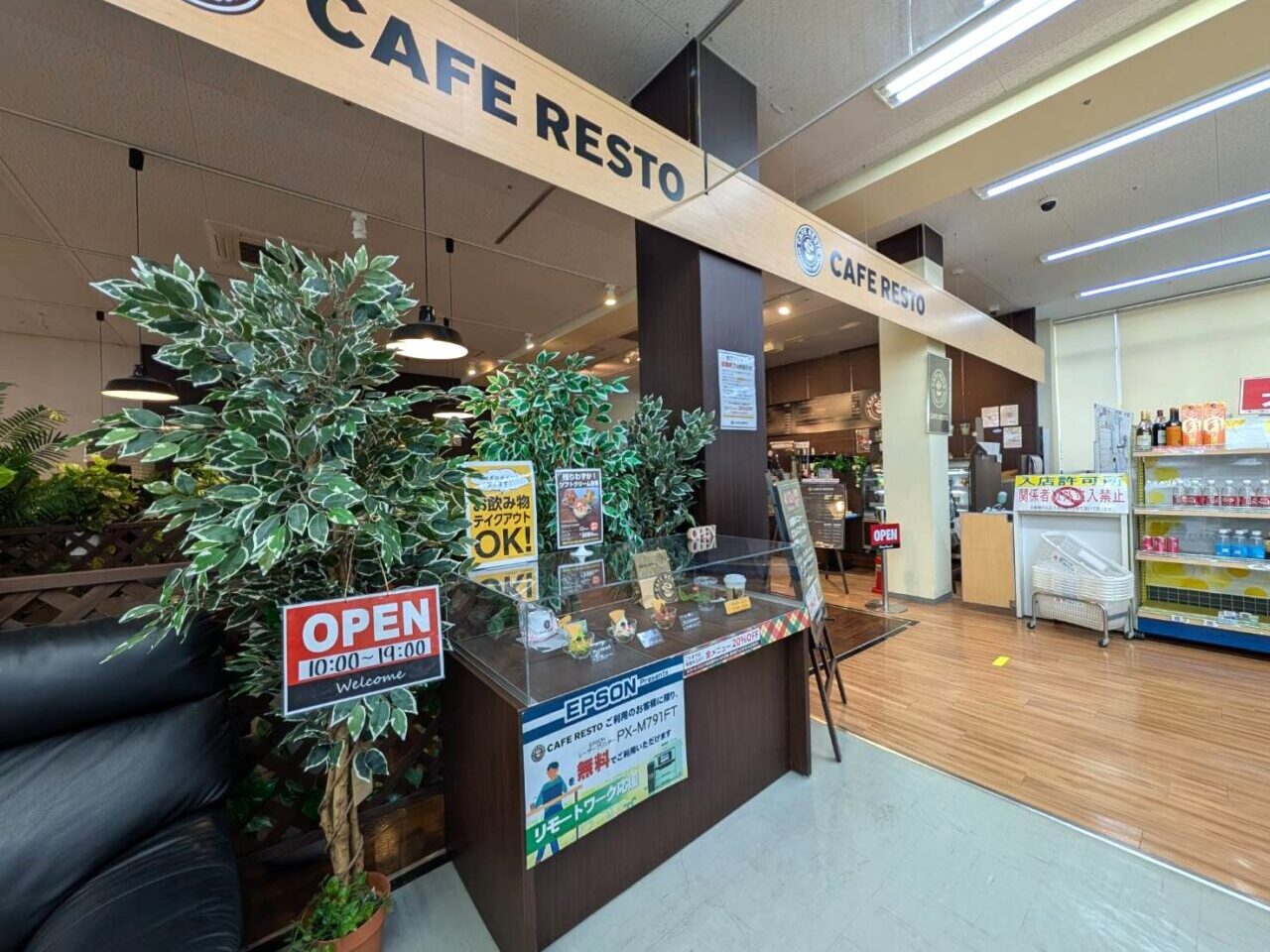 Gemini の回答 商業施設内にあるカフェ「CAFE RESTO」の入り口。木目調の看板、メニュー見本が並ぶカウンター、観葉植物があり、営業時間は10:00〜19:00と掲示されています。