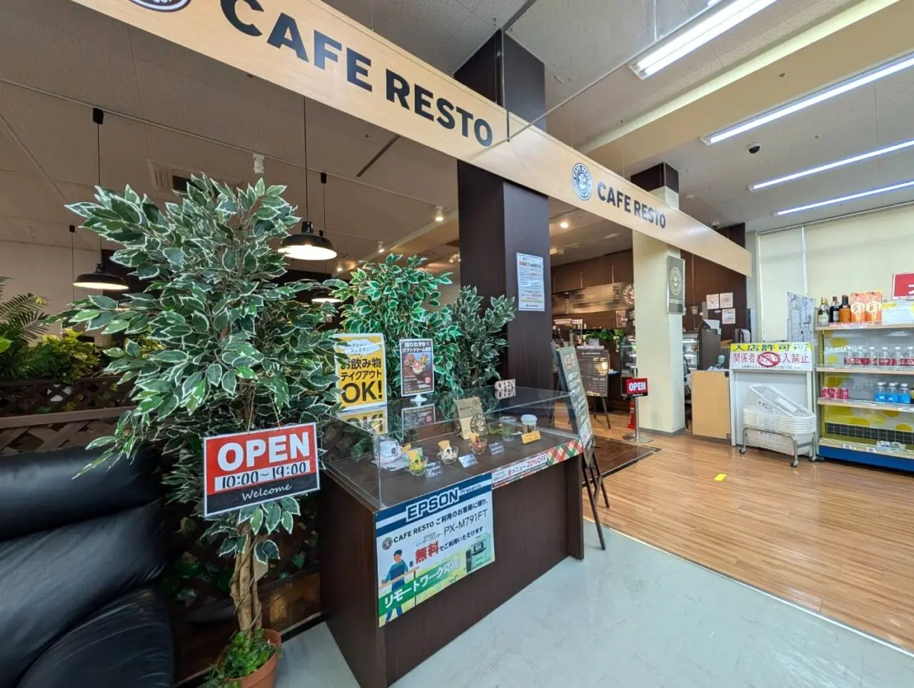 Gemini の回答
商業施設内にあるカフェ「CAFE RESTO」の入り口。木目調の看板、メニュー見本が並ぶカウンター、観葉植物があり、営業時間は10:00〜19:00と掲示されています。
